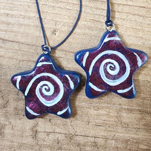 May include: Two star-shaped pendants with blue edges and a swirling white design on a burgundy background. The pendants are strung on blue cords. The pendants are on a wooden surface.