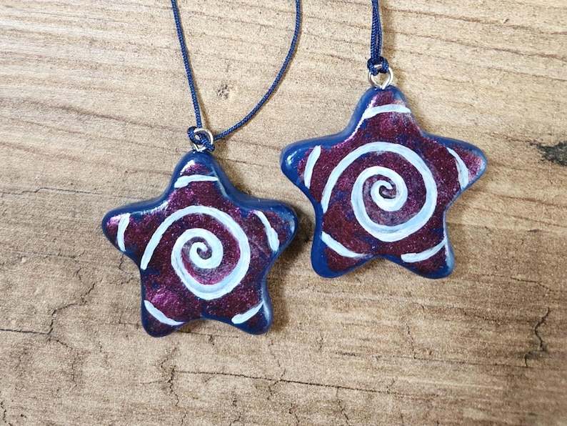 May include: Two star-shaped pendants with blue edges and a swirling white design on a burgundy background. The pendants are strung on blue cords. The pendants are on a wooden surface.