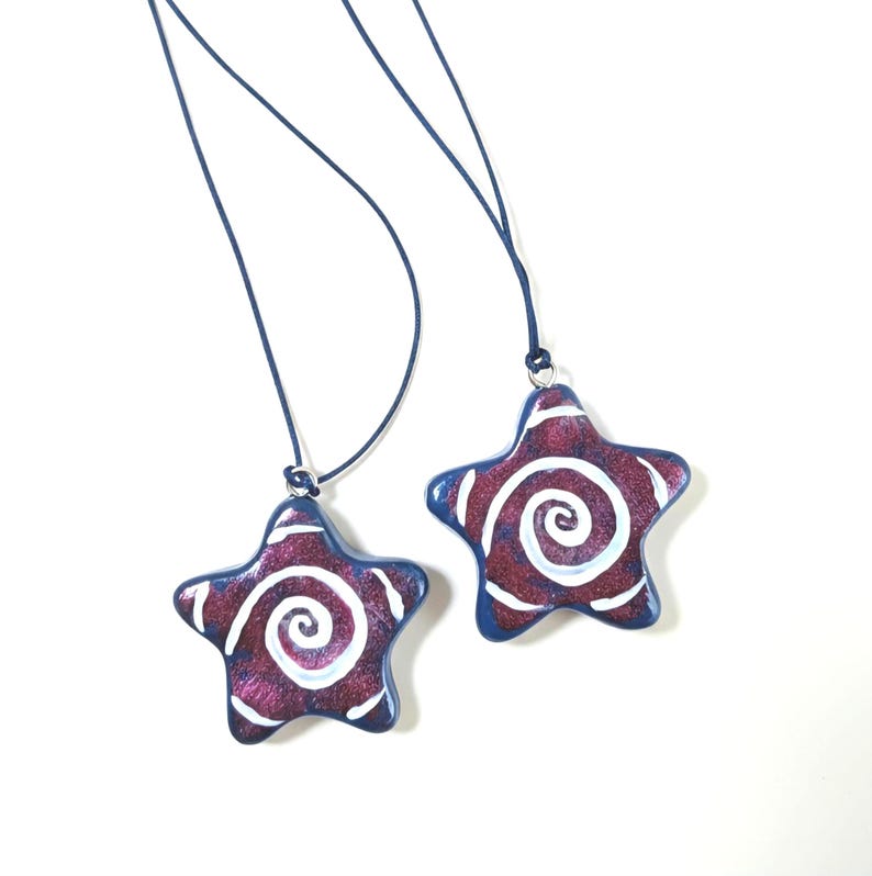 May include: Two star-shaped pendants with a swirling white design on a purple background. The pendants have a blue border and are suspended from a dark blue cord. The background is white.