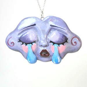May include: A pendant shaped like a stylized purple face with closed eyes, long black eyelashes, and pink cheeks. Two bright blue teardrops fall from the eyes, and the face has dark red lips. The pendant hangs from a thin silver-colored metal chain.