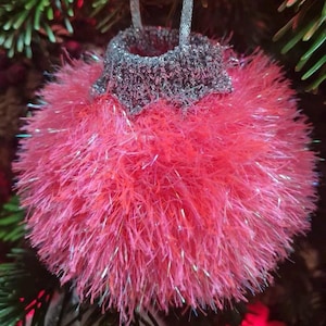 Puede incluir: Un adorno navideño de color rosa y peludo con detalles de purpurina plateada. El adorno es redondo y tiene un lazo en la parte superior para colgarlo.