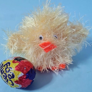 May include: A handmade, fluffy yellow chick with an orange beak and feet, and googly eyes. Next to the chick is a foil-wrapped chocolate egg with colorful packaging. The background is a light blue.
