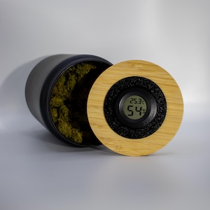 Könnte beinhalten: Ein schwarzer zylindrischer Behälter mit einem Holzdeckel. Der Deckel hat eine Digitalanzeige mit 25,3°C und 54%. Der Behälter ist geöffnet und zeigt grünen Inhalt. Der Deckel ist aus hellbraunem Holz mit schwarzem Rand.