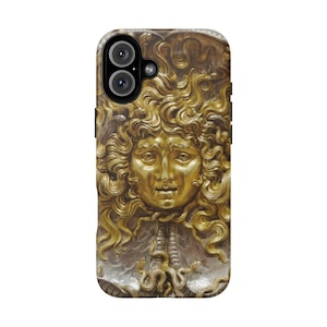 Op de afbeelding: Een telefoonhoesje met een gouden Medusa-hoofdontwerp. De hoes heeft een gedetailleerd reliëf van Medusa's gezicht met slangen als haar. De hoes is goud- en zilverkleurig, met een zwarte cameralens.
