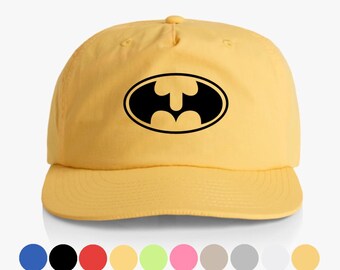Könnte beinhalten: Gelbe Baseballkappe mit schwarzem Batman-Logo in einem Oval. Die Kappe hat einen gebogenen Schirm und einen Stoffriemenverschluss. Eine Reihe von Farboptionen wird darunter angezeigt.