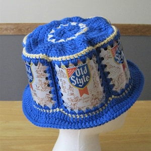 May include: A blue crocheted hat with a white crocheted brim. The hat is made from recycled beer cans with the Old Style beer logo.