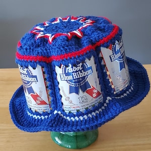 May include: A handmade blue crochet bucket hat with red and white accents. The hat features three Pabst Blue Ribbon beer cans incorporated into the design. The hat is a unique and quirky accessory.