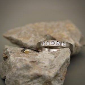 Può includere: Una fascia d'argento con una fila di piccoli diamanti trasparenti incastonati nel metallo. L'anello è appoggiato su un pezzo di roccia grigia ruvida.