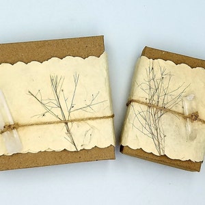 Pu&ograve; includere: Due piccole scatole rettangolari avvolte in carta marrone con un bordo decorativo a festoni. Le scatole sono legate con spago e decorate con fiori secchi e cristalli di quarzo trasparenti.