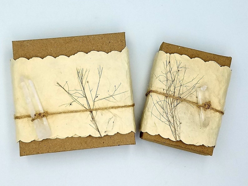 Pu&ograve; includere: Due piccole scatole rettangolari avvolte in carta marrone con un bordo decorativo a festoni. Le scatole sono legate con spago e decorate con fiori secchi e cristalli di quarzo trasparenti.