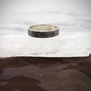 Könnte beinhalten: Ein silberner Bandring mit einem strukturierten schwarzen Design. Der Ring liegt auf einer weißen Oberfläche.