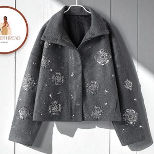 Pode incluir: Casaco de lã cinzento com gola alta e enfeites de lantejoulas prateadas em padrão de floco de neve. O casaco tem fecho de correr e está pendurado num cabide branco. O logótipo da marca "TRENDTHREAD" está num círculo no canto superior esquerdo.