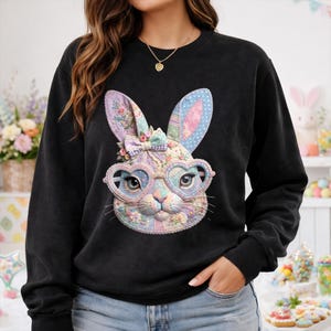 May include: Black sweatshirt featuring a colorful Easter bunny face with heart-shaped glasses. The bunny has floral and patchwork details, with a bow and polka dot accents on the ears. A charming and festive design for the spring season.