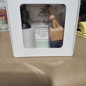 May include: A white box with a clear window showing a car air freshener kit. The kit includes a small bottle of essential oil, a wooden diffuser with a string, and a card that says "Car Air Diffuser".