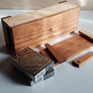 May include: A wooden box with a slot for holding a deck of cards. The box is made of two different types of wood, one light and one dark. The box is open and a deck of Bicycle playing cards is sitting on top of it. The cards have a black and gold design.