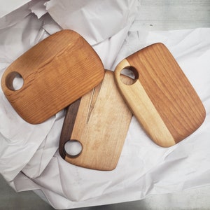 May include: Three wooden cutting boards with different wood grain patterns and a hole in the top. The boards are arranged on a white surface.