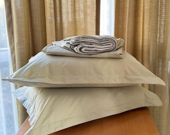 May include: A stack of three light green pillowcases on a wooden surface. The pillowcases are folded and have a subtle texture. The background is a set of beige curtains.