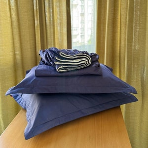 May include: A stack of blue pillowcases and a blue pillow on a wooden table. The pillowcases are folded and the pillow is fluffed. The table is in front of a window with yellow curtains.