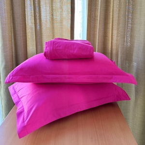 May include: A set of bright pink bed linens, including two pillowcases and a folded sheet, are arranged on a wooden surface. The linens are a vibrant fuchsia colour, contrasting with the neutral-toned curtains in the background.