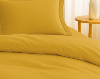 May include: Mustard yellow bedding set featuring two pillowcases with a contrasting border and a duvet cover. The set is displayed on a bed with a rounded headboard. A small side table with a white vase and greenery is visible.