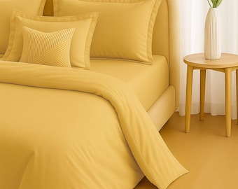 May include: A bed with yellow bedding, including a duvet, fitted sheet, and pillows. A small decorative pillow with a textured design is visible. A white vase with green leaves sits on a small wooden side table next to the bed.