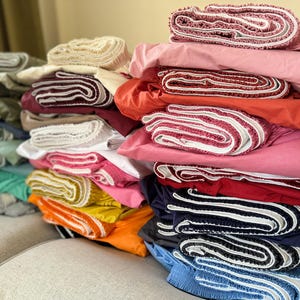 May include: A stack of colorful fabric rolls with white trim. The fabric rolls are folded and stacked on top of each other. The fabric rolls are in various colors, including red, pink, blue, green, yellow, and orange.