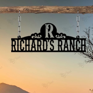 Puede incluir: Un letrero de metal negro con el texto "RICHARD'S RANCH" y la letra "R" en un círculo sobre el texto. El letrero está colgado de una viga de madera con cadenas.