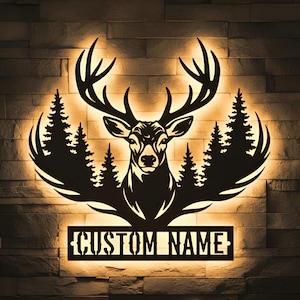 Puede incluir: Silueta metálica negra de una cabeza de ciervo con grandes astas, rodeada de pinos. El diseño incluye un área rectangular en la parte inferior con las palabras "CUSTOM NAME" recortadas. La obra de arte está iluminada contra una pared de ladrillos.