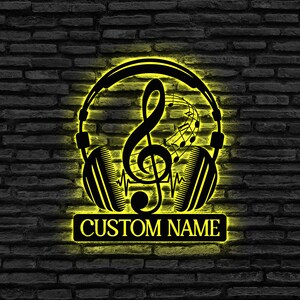 Könnte beinhalten: Schwarze Metall-Wandkunst mit einem musikalischen Thema. Das Design umfasst einen Notenschlüssel, Noten und Kopfhörer, die alle mit einem gelben Schein beleuchtet sind. Der Text "CUSTOM NAME" wird auf einem rechteckigen Schild angezeigt.
