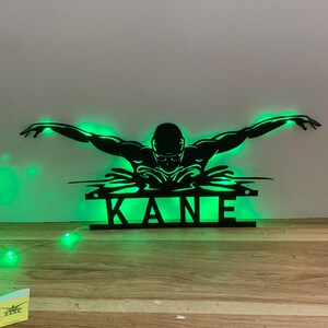 Peut inclure: Une silhouette noire en métal d'un nageur en papillon avec le nom "KANE" en dessous. La silhouette est éclairée par des lumières LED vertes.
