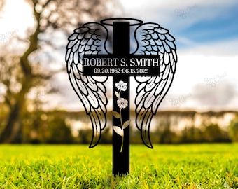 May include: Black metal memorial cross with angel wings, a halo, and a floral accent. The cross features the name "ROBERT S. SMITH" and dates "09.20.1962-06.15.2025". The cross is set in a grassy field.