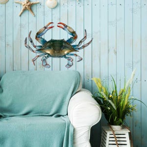 May include: A blue crab wall decoration with realistic details. The crab is made of metal and has a blue, green and orange colour scheme. It is mounted on a light blue wooden wall with a white sofa and a potted plant in the background.