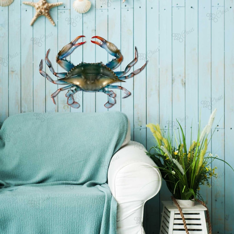 May include: A blue crab wall decoration with realistic details. The crab is made of metal and has a blue, green and orange colour scheme. It is mounted on a light blue wooden wall with a white sofa and a potted plant in the background.
