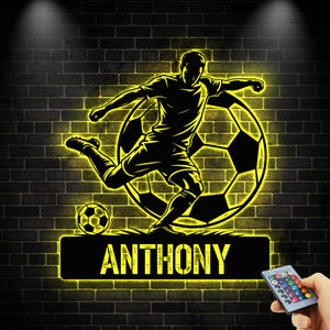 May include: A black and yellow illuminated soccer-themed wall art. The design features a soccer player kicking a ball, with the name "ANTHONY" below. The background is a brick wall.