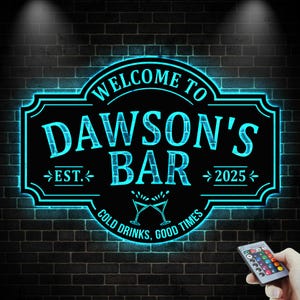 May include: A black, illuminated sign with the text "WELCOME TO DAWSON'S BAR" in a teal font. The sign includes "EST. 2025" and "COLD DRINKS, GOOD TIMES." The sign is mounted on a dark brick wall.
