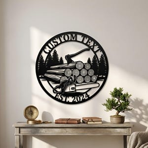 Pode incluir: Uma placa de parede circular em metal preto com um design recortado de uma pilha de troncos, um machado, uma motosserra e pinheiros. O texto CUSTOM TEXT está arqueado no topo e EST. 2024 na parte inferior da moldura circular.