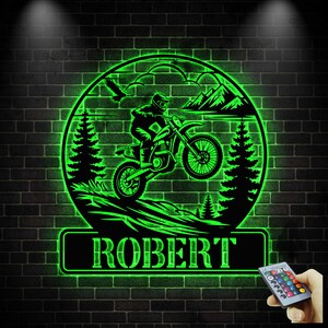 Może przedstawiać: Neonowo-zielone, okrągłe dzieło sztuki ściennej przedstawiające motocyklistę, góry i drzewa. Imię "ROBERT" jest wyświetlane na prostokątnej tabliczce poniżej. Grafika jest zamontowana na ciemnej ceglanej ścianie.