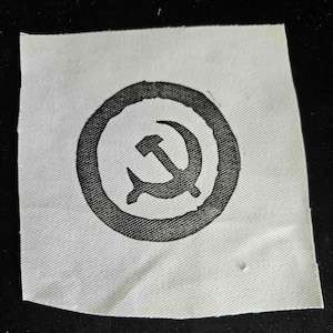 Könnte beinhalten: Ein quadratischer, cremeweißer Stoffaufnäher mit einem schwarzen Aufdruck. Das Motiv besteht aus einem soliden schwarzen Kreis, der ein stilisiertes Hammer-und-Sichel-Symbol in der Mitte enthält. Der Stoff weist eine sichtbare diagonale Webstruktur auf.