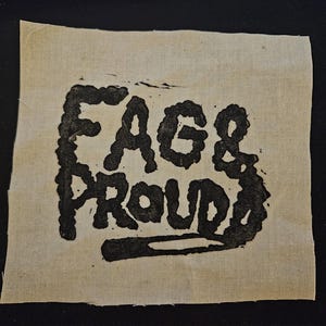 Könnte beinhalten: Ein quadratischer Aufnäher aus cremefarbenem Stoff mit dem schwarzen Schriftzug "FAGS & PROUD". Die Buchstaben sind dick und stilisiert, mit einem handgefertigten Aussehen. Der Stoff hat eine sichtbare Textur und ausgefranste Kanten.