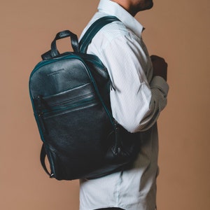 May include: A black leather backpack with green trim and a zipper closure. The backpack is being worn by a person wearing a white striped shirt and black pants.