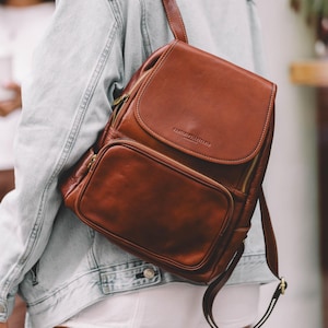 Peut inclure: Un sac à dos en cuir marron avec des fermetures éclair dorées et un petit logo sur le devant. Le sac à dos est porté par une personne portant une veste en jean bleu clair.