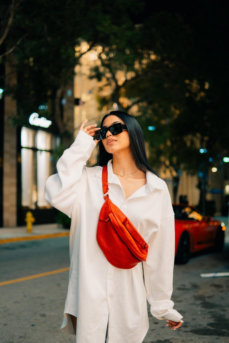Trendy red leather hip bag worn crossbody by a stylish woman in an urban setting. This modern leather bag combines luxury and functionality, perfect as a fashion-forward festival bag, everyday carry, or stylish crossbody for travel and city life.