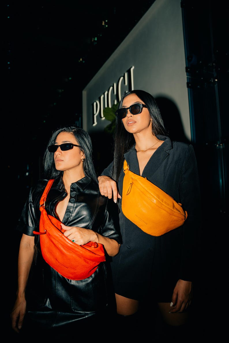 Two women wearing modern leather hip bags, one in vibrant red and the other in bold yellow. Perfect for festivals and everyday carry, these stylish crossbody fanny packs offer a trendy way to carry essentials while adding a pop of color.