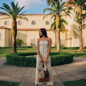 Woman standing outdoors in front of a beautiful building with palm trees, holding a modern leather handbag.