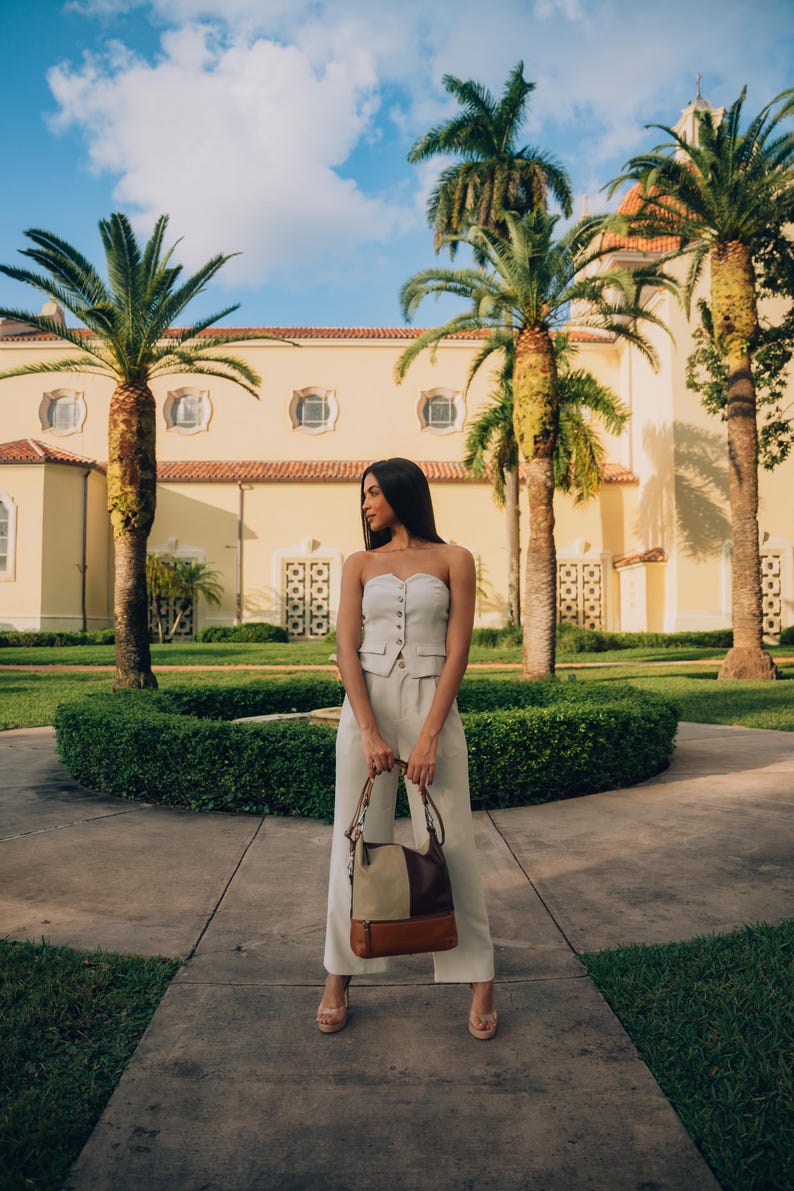 Woman standing outdoors in front of a beautiful building with palm trees, holding a modern leather handbag.