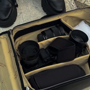 Peut inclure: Sac d'appareil photo en cuir noir ouvert, révélant des objectifs et accessoires noirs. Le sac est compartimenté. Le sac est posé sur une surface en pierre.