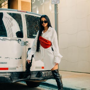 A stylish woman poses outdoors with a red leather fanny pack worn crossbody, paired with an oversized white shirt and thigh-high boots, giving a bold, fashion-forward look.