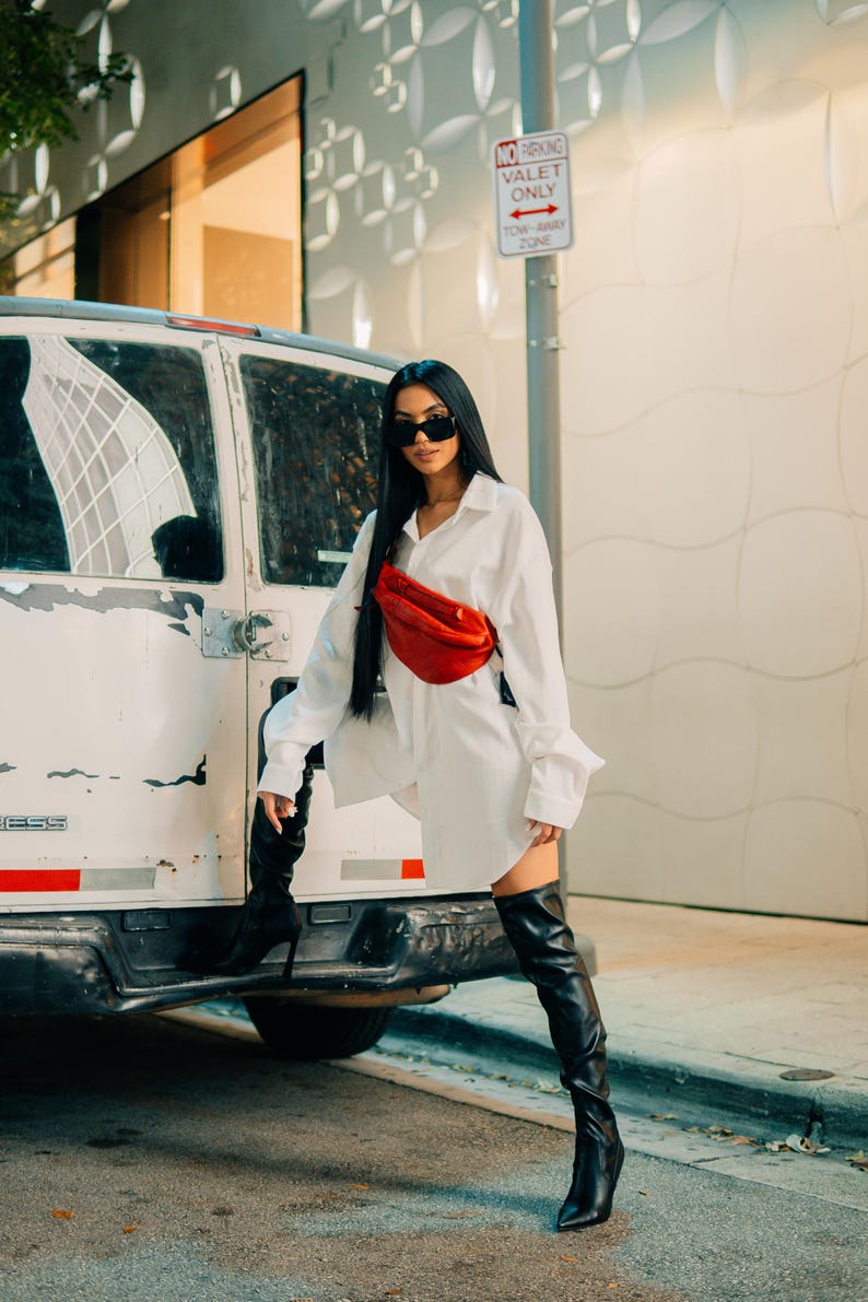 A stylish woman poses outdoors with a red leather fanny pack worn crossbody, paired with an oversized white shirt and thigh-high boots, giving a bold, fashion-forward look.