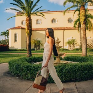 Woman walking through a sunny garden with palm trees, carrying a leather tote bag in neutral shades