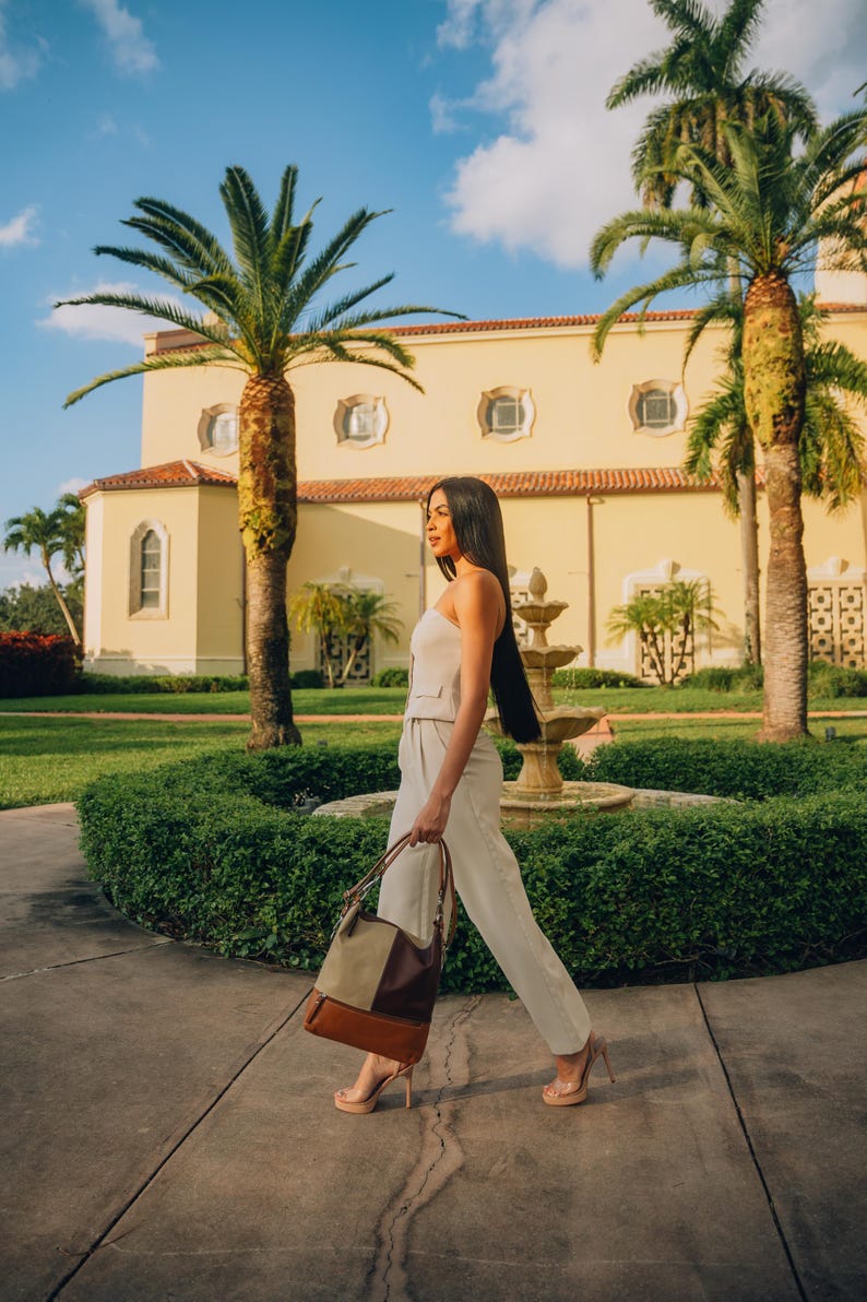 Woman walking through a sunny garden with palm trees, carrying a leather tote bag in neutral shades